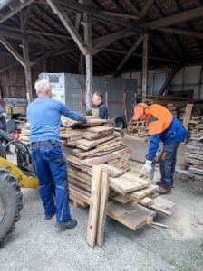 Praktischer Einsatz Draußen Ein Radlader hat mit seiner Gabel einen großen Bretterstapel geladen. Drei Männer arbeiten daran: 2 Männer verladen das Holz, 1 Mann sägt die Bretter mit einer Motorsäge kleiner