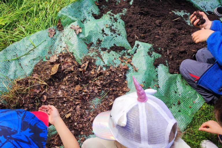 Im Garten Gehts Rund Kinder sind über einen Haufen feinen und einen Haufen groben Kompost gebeugt und untersuchen ihn. Ein Kind hat eine Assel in der Hand, ein anderes die feine Erde