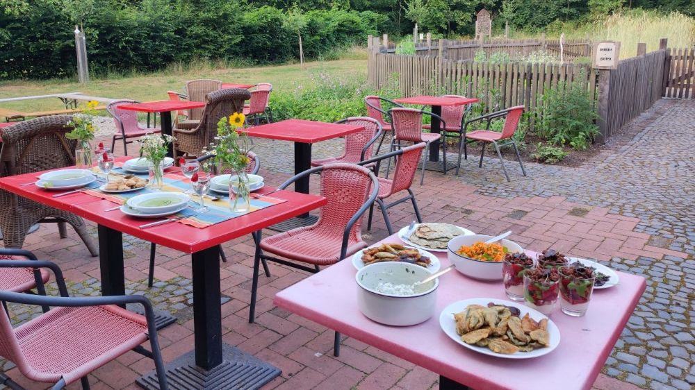 Im Ramsbrocker Garten steht eine gedeckte Tafel . Auf einer längeren Tischreihe stehen Suppenteller mit Kräutersuppe, auf einem Beistelltisch die speisen der anderen Gänge. Der Tisch ist mit Vasen voller Wildblumen dekoriert