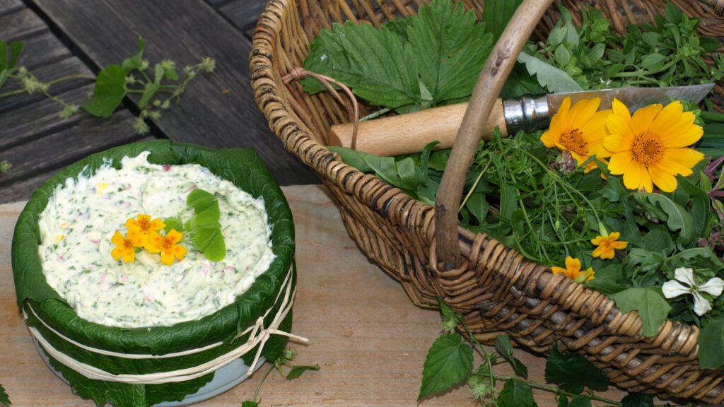 Auf einem Holzbrett steht ein Schälchen mit Wildkräuteraufstrich, garniert mit gelben Blüten. Daneben steht ein geflochtener Weidenkorb, in dem weitere Wildkräuter liegen, gelbe Ringelblumen leuchten im Korb und ein Taschenmesser liegt auf den Kräutern